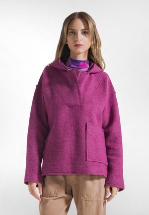 Jonge vrouw met lang haar, gekleed in een paarse capuchonsweater, beige broek en een kleurrijke sjaal, staand tegen een effen achtergrond.