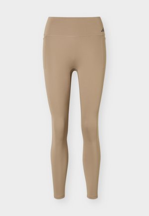 Μπεζ κολάν ψηλόμεσο, μακρύ έως τον αστράγαλο, με πλαϊνές τσέπες και λογότυπο Adidas στη ζώνη μέσης, σχεδιασμένο για αθλητική χρήση.