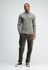 Grijze gebreide ritsvest met geribbelde manchetten, gecombineerd met donkerbruine broek en zwarte sneakers met witte zolen. Subtiele logodetail op de borst.