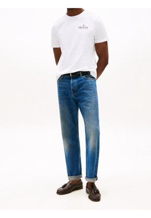 Mand iført hvid Tommy Hilfiger t-shirt, blå oprullede jeans, sort bælte og mørkebrune tassel loafers, stående på hvid baggrund.