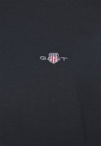 Tela azul marino con un pequeño logo bordado de GANT que presenta un escudo con estrellas y franjas rojas en hilo blanco.