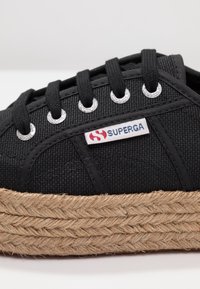 Zapatillas de lona negras con suela de yute trenzado. Presentan ojales plateados y una etiqueta blanca con una marca en rojo y azul. Cierre de cordones planos.