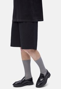 Piernas con pantalones cortos negros, calcetas grises hasta la mitad de la pierna y mocasines de cuero negro contra un fondo claro y liso.