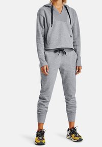 Donna che indossa un hoodie grigio di Under Armour e pantaloni da jogging con lacci neri e scarpe da corsa multicolori, in piedi contro uno sfondo bianco.
