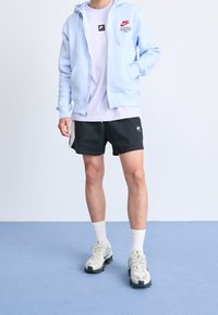 Sudadera con cremallera de color azul claro con logo, combinada con una camiseta lavanda y pantalones cortos negros. Zapatillas beige y verdes con suela texturizada y calcetines blancos.