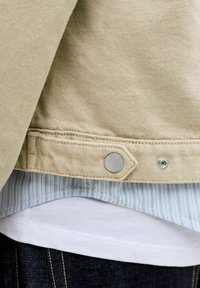 Beige Jeansjacke mit Metall-Druckknöpfen über einem blau gestreiften Hemd und einem weißen Unterhemd, kombiniert mit dunklen Jeans.