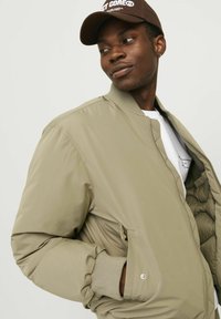 Veste bomber beige avec des poignets côtelés et fermeture éclair. Texture lisse et rembourrée, avec des poches latérales et une coupe décontractée.