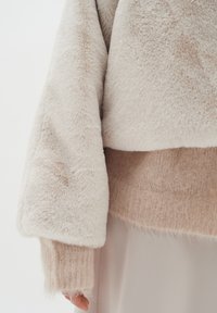 Beige kortärmad tröja med en mjuk, fluffig textur, breda ärmar och en lagerdesign, kompletterad med ett ljusare inre material.