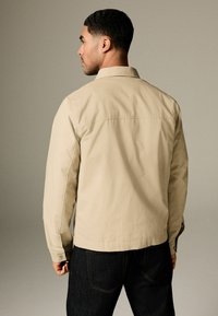 Veste décontractée beige clair avec un col, des manches longues et des poignets boutonnés. Portée sur un jeans foncé, vue de dos sur un fond neutre.