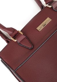 Sac à main en cuir bordeaux avec une surface texturée, plaque de marque à tonalité dorée et bretelles ajustables. Présente des accents cousus et une forme rectangulaire.