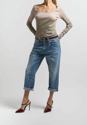 Kvinde iført beige top med rund hals og lange ærmer, højtsiddende, opkrogede blå jeans og bordeauxfarvede spidse højhælede sko i stående positur.