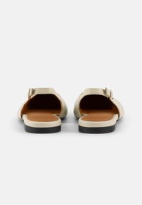 Sandalias abiertas de color beige claro, hechas de un material suave, con una plantilla marrón, suela plana negra y un pequeño detalle de hebilla metálica.