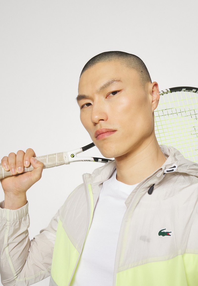 Lichte, beige jas met neongele accenten. Model houdt een tennisracket vast, met een gestructureerde grip en een zwart frame.