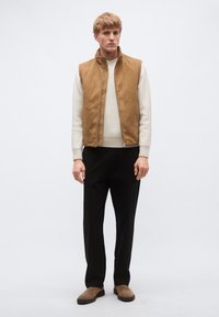 Tan antilop telovnik čez kremasto rebrasto pulover, s črnimi hlačami in rjavimi čevlji. Enostaven, krojen dizajn z detajli z zadrgo.