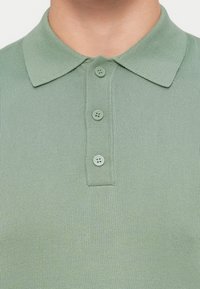 Κοντινό πλάνο ενός ατόμου που φοράει ένα ανοιχτοπράσινο πλεκτό πόλο με γιακά και τρία κουμπιά στο ίδιο πράσινο χρώμα κλεισμένα.