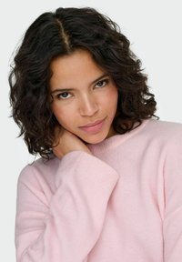 Light pink, textured sweater with a relaxed fit and ribbed cuffs. Model has dark, curly hair with a natural sheen, hand resting on her neck.