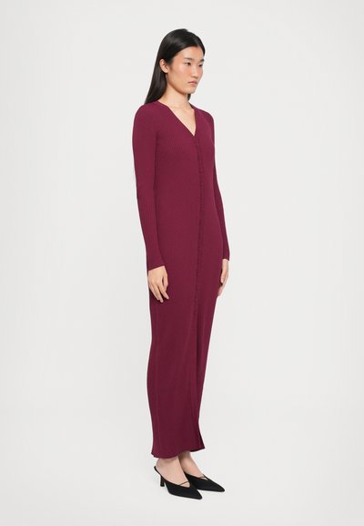 Long maroon ribbed knit dress with a V-neck, full-length sleeves, and button closure, paired with black pointed-toe heels.