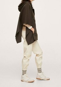 Poncho à capuche vert foncé à coupe décontractée, associé à des joggers crème et des baskets montantes blanches, avec des accents beige et des poignets côtelés.