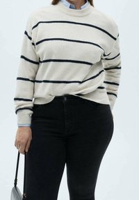 Femme portant un pull blanc à rayures horizontales noires, un pantalon noir, tenant un sac à main noir sur un fond uni.