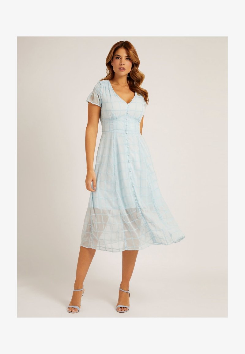 Femme portant une robe midi bleu clair à manches courtes boutonnée avec motif à carreaux et des talons à bride à la cheville assortis, debout sur un fond clair.