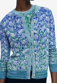 Neulottu neuletakki sinivihreällä kukkakuviolla. Siinä on teksturoitu kaulus ja napitettava suljenta, resorit hihansuissa ja helmassa.