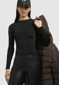 Top nero a maniche lunghe, elegante e aderente. Leggings neri lucidi. Giacca a vento trapuntata marrone scuro tenuta sulla spalla. Cappellino nero con logo.