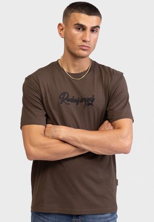 Άνδρας με κοντά μαλλιά, φορώντας καφέ μπλουζάκι "Redefined Root", χρυσή αλυσίδα και ασημένιο βραχιόλι, στέκεται με σταυρωμένα τα χέρια.