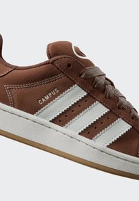 Brown suede sneakers with white leather stripes, metal eyelets, and a textured white rubber sole. Features a logo on the tongue.