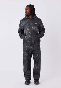 Schwarzer Tracksuit mit Batikmuster, bestehend aus einer zipbaren Kapuzenjacke und Cargo-Hosen. Verfügt über Taschen und ein Logo auf der Brust. Strukturiertes Finish.