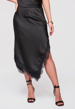Kobieta nosząca czarną satynową spódnicę midi z koronkową lamówką oraz czarne sandały na wysokim obcasie z paskami, stojąca na jednolitym szarym tle.