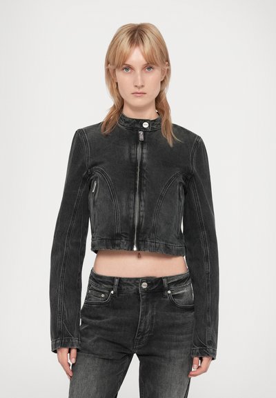 Personne blonde portant une veste en denim noire courte avec fermeture éclair et col montant, associée à un jean en denim noir taille haute assorti.
