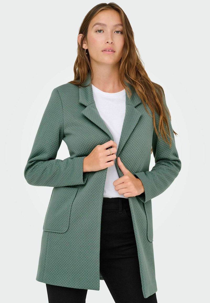 Manteau vert texturé avec un col cranté, design à un seul bouton et deux poches avant, porté sur une chemise blanche et un pantalon noir.