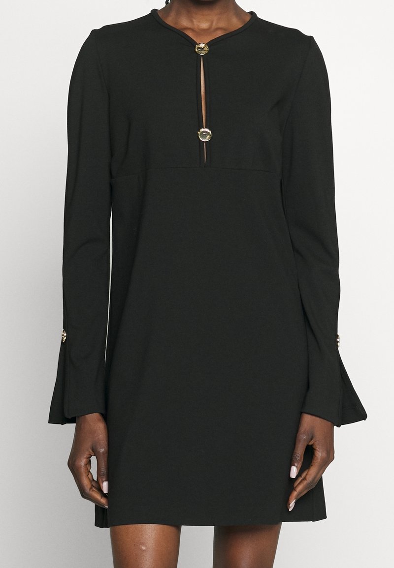 Robe noire à manches longues avec ouverture en forme de trou sur le devant et deux boutons dorés décoratifs, poignets évasés avec détails de boutons assortis.