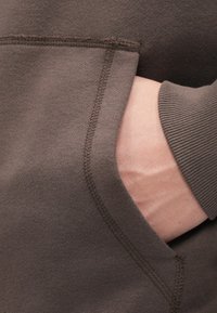 Tessuto di felpa marrone con una texture morbida, dotato di una tasca frontale e polsini a coste. Dettagli di cucitura lungo le cuciture.