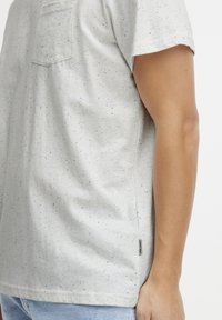 Tricou din bumbac gri cu pete, prevăzut cu un buzunar pe piept și o etichetă pe lateral. Stofful pare moale, cu o croială lejeră și mâneci scurte.