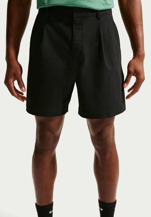 Mann trägt schwarze plissierte Shorts und schwarze Nike-Socken und steht vor einfachem Hintergrund, die Hände entspannt an den Seiten.
