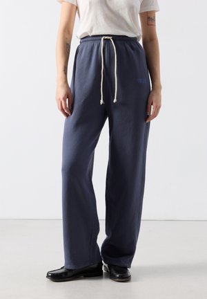 Persoon draagt marineblauwe losse sweatpants met witte trekkoord, een wit shirt met korte mouwen en zwarte loafers op een lichte vloer.