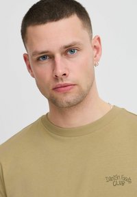 Alyvuogių spalvos medvilninė marškinė su apvaliu kaklu ir trumpomis rankovėmis, ant kairiojo krūtinės pusėje siuvinėta tamsiai žaliai "Danish Foodie Club".