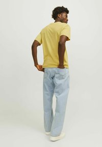 Camiseta de algodón amarilla con mangas cortas, combinada con jeans de mezclilla azul claro. El modelo está de espaldas, con las manos en los bolsillos, usando zapatos blancos.