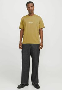 Olive green cotton t-shirt with short sleeves and central white logo, paired with dark denim wide-leg pants and black sturdy boots.