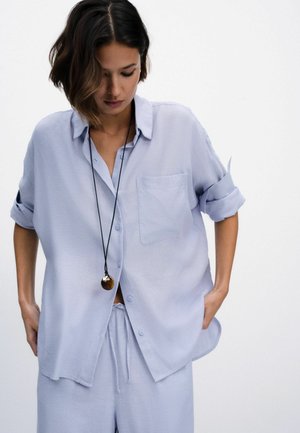 Sieviete tērpusies vaļīgā gaiši zilā kreklā ar uzrullētām piedurknēm, atbilstošās biksēs un garā kaklarotā ar apaļu piekariņu, skata vērsts lejup.