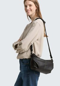 Schwarze Leder-Schultertasche mit strukturiertem Finish, geschwungener Form und kontrastierenden Nähten, die an der Schulter einer Person gegen einen hellen Hintergrund getragen wird.