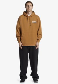 Felpa in pile marrone con tasca a kanga frontale e cappuccio con cordino. Abbinata a pantaloni neri larghi e scarpe nere. Presenta una toppa con logo.