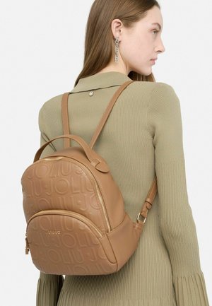 Zaino in pelle marrone chiaro con logo embossato, forma arrotondata, doppia cerniera e spallacci regolabili, indossato sulla schiena di una persona con una maglietta a coste.