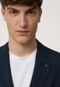 Navy blazer made of textured fabric with a blue dog embroidery on the lapel. Worn over a plain white T-shirt.