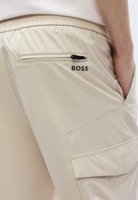 Μπεζ παντελόνι φόρμας με φερμουάρ στην πίσω τσέπη, εκτυπωμένο λογότυπο κοντά στην τσέπη και λείο, ελαφρύ ύφασμα με διακριτική γυαλάδα.