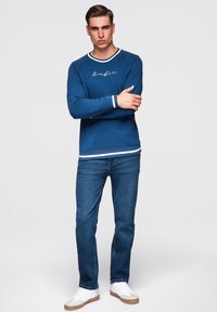 Mladý muž stojí se zkříženýma rukama, oblečený v modrém svetru s bílými pruhy, modrých džínách a bílých teniskách na jednotném pozadí.