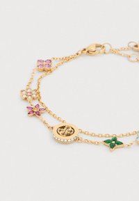 Βραχιόλι σε χρυσό χρώμα με πολλαπλά γούρια, συμπεριλαμβανομένων λουλουδιών και στρογγυλού σχεδίου, με ροζ, πράσινες και διάφανες πολύτιμες πέτρες. Ρυθμιζόμενο κούμπωμα.