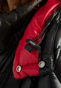 Giacca puffer nera e rossa con una texture lucida, dotata di una corda di regolazione nera e bottoni a pressione argentati per la chiusura.