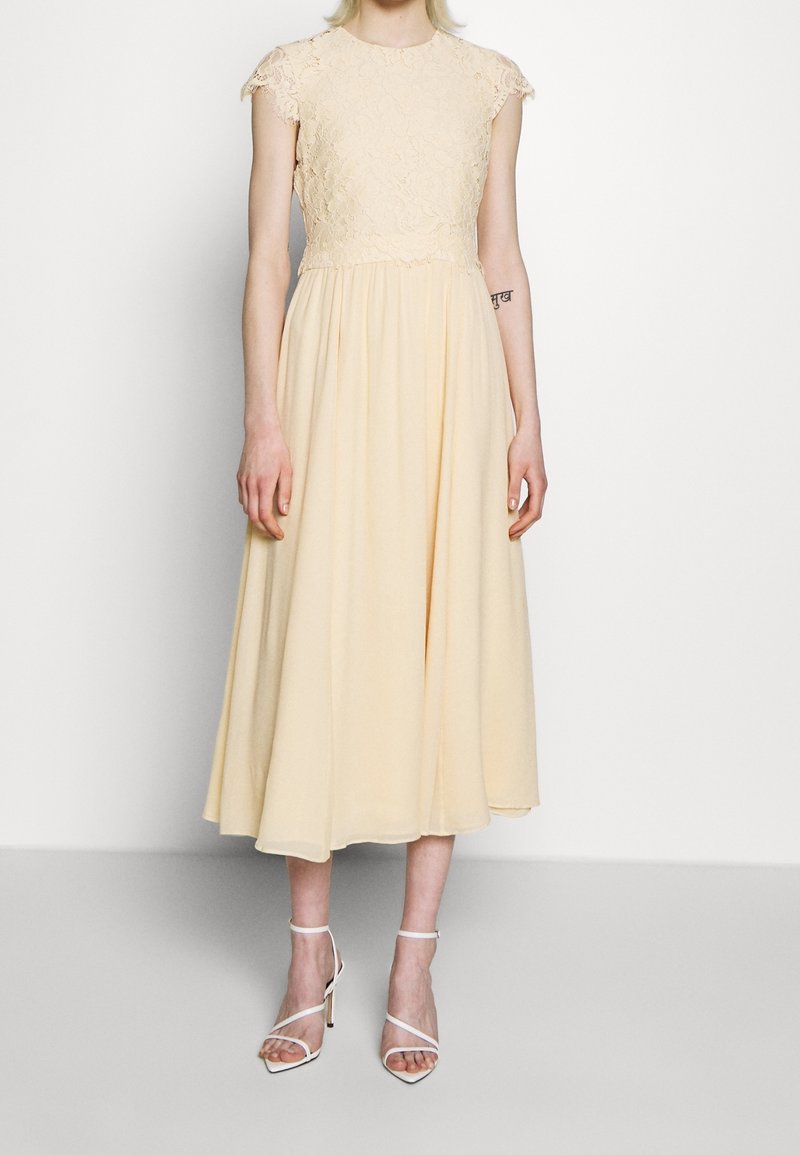 Kvinna klädd i en ljusgul spetsklänning med en svepande kjol och vita remmar med hög klack, som står mot en enkel vit bakgrund.